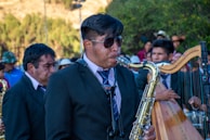 A man wearing sunglasses and a dark suit plays the saxophone in an outdoor setting with several other musicians and people around. The scene is lively with a mixture of musical instruments, including a harp, visible in the background. The ensemble seems to be performing, suggesting a festive or ceremonial atmosphere.