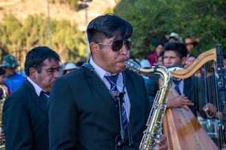 A man wearing sunglasses and a dark suit plays the saxophone in an outdoor setting with several other musicians and people around. The scene is lively with a mixture of musical instruments, including a harp, visible in the background. The ensemble seems to be performing, suggesting a festive or ceremonial atmosphere.