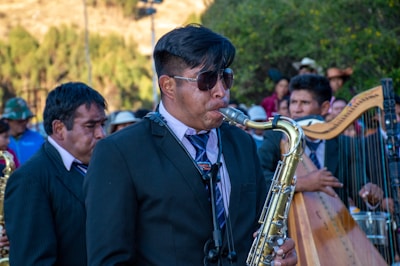 A man wearing sunglasses and a dark suit plays the saxophone in an outdoor setting with several other musicians and people around. The scene is lively with a mixture of musical instruments, including a harp, visible in the background. The ensemble seems to be performing, suggesting a festive or ceremonial atmosphere.