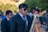 A man wearing sunglasses and a dark suit plays the saxophone in an outdoor setting with several other musicians and people around. The scene is lively with a mixture of musical instruments, including a harp, visible in the background. The ensemble seems to be performing, suggesting a festive or ceremonial atmosphere.