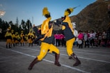 Two individuals are performing a dynamic dance or ritual in colorful costumes, featuring bright yellow and black garments with prominent feathered headpieces. In the background, several similarly dressed participants stand in line, while a group of people dressed in pink observes the scene. The setting appears to be outdoors, with a mountainous landscape providing the backdrop.