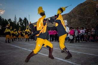 Two individuals are performing a dynamic dance or ritual in colorful costumes, featuring bright yellow and black garments with prominent feathered headpieces. In the background, several similarly dressed participants stand in line, while a group of people dressed in pink observes the scene. The setting appears to be outdoors, with a mountainous landscape providing the backdrop.