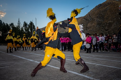 Two individuals are performing a dynamic dance or ritual in colorful costumes, featuring bright yellow and black garments with prominent feathered headpieces. In the background, several similarly dressed participants stand in line, while a group of people dressed in pink observes the scene. The setting appears to be outdoors, with a mountainous landscape providing the backdrop.