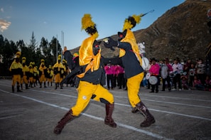 Two individuals are performing a dynamic dance or ritual in colorful costumes, featuring bright yellow and black garments with prominent feathered headpieces. In the background, several similarly dressed participants stand in line, while a group of people dressed in pink observes the scene. The setting appears to be outdoors, with a mountainous landscape providing the backdrop.