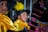 A young boy in traditional attire, featuring a yellow scarf and a black hat adorned with a vibrant yellow feather. He is among other children wearing colorful garments. An adult, possibly a woman, stands behind them wearing an intricately patterned outfit.