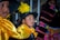 A young boy in traditional attire, featuring a yellow scarf and a black hat adorned with a vibrant yellow feather. He is among other children wearing colorful garments. An adult, possibly a woman, stands behind them wearing an intricately patterned outfit.