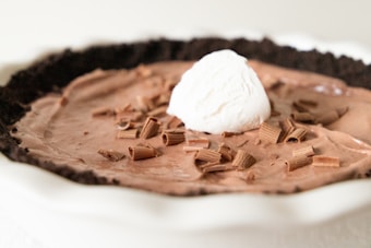 A creamy chocolate dessert pie topped with chocolate shavings and a dollop of whipped cream sits inside a dark crumb crust. The textures are smooth and fluffy, with a rich, indulgent appearance.