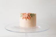 A small, elegantly decorated cake sits on a glass cake stand. The cake is adorned with a floral design featuring orange petals and green leaves, accentuated with tiny white pearl-like decorations on the top.