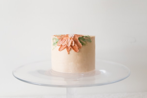 A small, elegantly decorated cake sits on a glass cake stand. The cake is adorned with a floral design featuring orange petals and green leaves, accentuated with tiny white pearl-like decorations on the top.