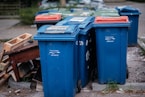 A row of blue recycling bins is lined up on the pavement, with some showing wear and tear. In the background, there are a few more bins with red and green lids. To the side, there are pieces of discarded wooden furniture stacked haphazardly.