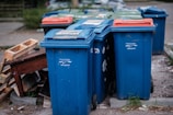 A row of blue recycling bins is lined up on the pavement, with some showing wear and tear. In the background, there are a few more bins with red and green lids. To the side, there are pieces of discarded wooden furniture stacked haphazardly.