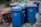 A row of blue recycling bins is lined up on the pavement, with some showing wear and tear. In the background, there are a few more bins with red and green lids. To the side, there are pieces of discarded wooden furniture stacked haphazardly.