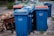 A row of blue recycling bins is lined up on the pavement, with some showing wear and tear. In the background, there are a few more bins with red and green lids. To the side, there are pieces of discarded wooden furniture stacked haphazardly.