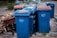 A row of blue recycling bins is lined up on the pavement, with some showing wear and tear. In the background, there are a few more bins with red and green lids. To the side, there are pieces of discarded wooden furniture stacked haphazardly.