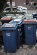 A row of several garbage bins in blue and green colors, with some having lids in different shades of orange. The bins are lined up along a street or sidewalk, partially surrounded by grass and concrete. Cars are parked in the background alongside residential buildings.