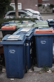 A row of several garbage bins in blue and green colors, with some having lids in different shades of orange. The bins are lined up along a street or sidewalk, partially surrounded by grass and concrete. Cars are parked in the background alongside residential buildings.