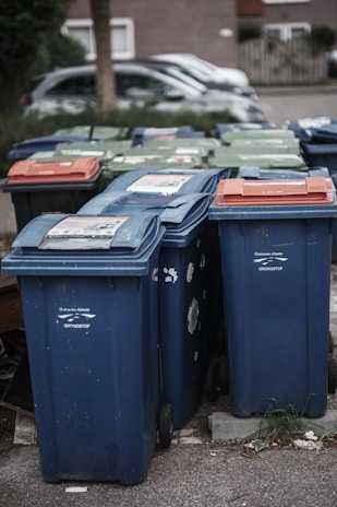 A row of several garbage bins in blue and green colors, with some having lids in different shades of orange. The bins are lined up along a street or sidewalk, partially surrounded by grass and concrete. Cars are parked in the background alongside residential buildings.