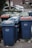 A row of several garbage bins in blue and green colors, with some having lids in different shades of orange. The bins are lined up along a street or sidewalk, partially surrounded by grass and concrete. Cars are parked in the background alongside residential buildings.