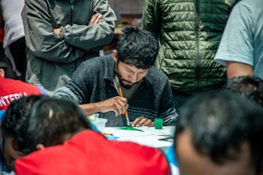A man intensely focuses on painting at a table, surrounded by other people. He is holding a brush and there are paint supplies nearby. Some individuals stand with arms crossed, wearing various jackets.