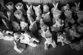 An assortment of unpainted plaster or ceramic figurines arranged closely together. The figures include various animals and cartoon-like characters with exaggerated features. The composition is in black and white, emphasizing the textures and details of the figurines, which are placed on a rough, textured surface.