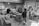 Several people are gathered in a workshop, focusing on a central wooden workbench surrounded by various tools, wood planks, and shelves filled with materials. The setting is industrial with a rough concrete floor and walls adorned with assorted papers and items.