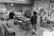 Several people are gathered in a workshop, focusing on a central wooden workbench surrounded by various tools, wood planks, and shelves filled with materials. The setting is industrial with a rough concrete floor and walls adorned with assorted papers and items.