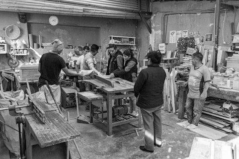 Several people are gathered in a workshop, focusing on a central wooden workbench surrounded by various tools, wood planks, and shelves filled with materials. The setting is industrial with a rough concrete floor and walls adorned with assorted papers and items.