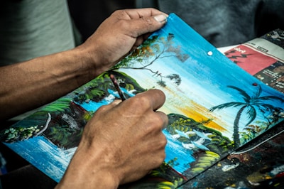 Hands of a person working on a detailed painting that depicts a landscape featuring a waterfall, palm trees, and a setting or rising sun casting a golden hue across the sky.