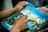 Hands of a person working on a detailed painting that depicts a landscape featuring a waterfall, palm trees, and a setting or rising sun casting a golden hue across the sky.
