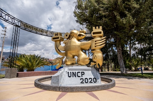 A large golden statue with animalistic features stands prominently on a pedestal labeled 'UNCP 2020' in front of a tree-lined campus area. The statue appears to be a stylized representation, holding various symbolic items in its hands. There is a sign above it that reads 'Universidad Nacional de Peru'. The background shows some buildings, trees, and a partly cloudy sky.