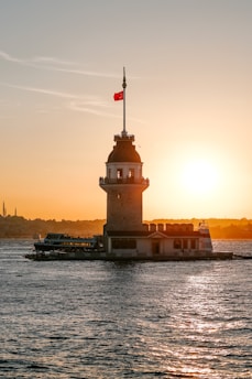 A historic tower stands majestically on a small islet, silhouetted against a vibrant sunset. The sky is painted with warm hues of orange and yellow, while the surrounding water reflects these colors. A Turkish flag flutters atop the tower. In the background, a faint outline of a cityscape is visible.
