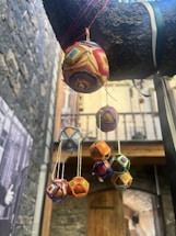 Colorful geometric hanging ornaments made from thread and wood hang from a branch. The background features a rustic stone wall and a wooden door under an archway.