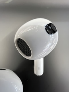 A close-up view of a white wireless earbud with a shiny surface and mesh details. The earbud features a small 'R' symbol indicating the right ear. The background is a smooth gradient of gray tones.