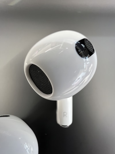 A close-up view of a white wireless earbud with a shiny surface and mesh details. The earbud features a small 'R' symbol indicating the right ear. The background is a smooth gradient of gray tones.