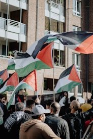 A group of people holding flags with black, white, green, and red colors are gathered in front of a brick building with balconies. Some individuals have raised their fists, and the flags are waving mid-air. The clothing suggests a variety of styles, indicating diverse participants.