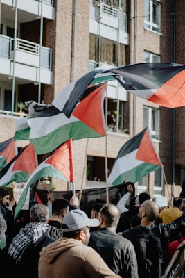 A group of people holding flags with black, white, green, and red colors are gathered in front of a brick building with balconies. Some individuals have raised their fists, and the flags are waving mid-air. The clothing suggests a variety of styles, indicating diverse participants.