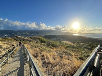 A scenic trail path with metal railings leads through a grassy landscape, overlooking a vast view of hills, a cityscape, and a bright sun setting over distant mountains. The sky is filled with scattered clouds, and the warm glow of the sun casts long shadows on the trail. Two people are walking along the path, enhancing the sense of scale and tranquility.