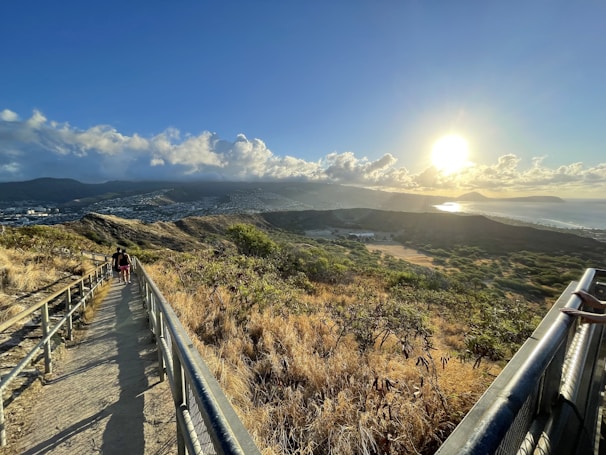 A scenic trail path with metal railings leads through a grassy landscape, overlooking a vast view of hills, a cityscape, and a bright sun setting over distant mountains. The sky is filled with scattered clouds, and the warm glow of the sun casts long shadows on the trail. Two people are walking along the path, enhancing the sense of scale and tranquility.