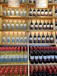 Shelves filled with rows of wine bottles, organized in an orderly fashion. The bottles have labels with blue and white designs, and the necks of the bottles are capped with red tops. Above the shelves, signs indicate different varieties and prices such as 'Tidal Bay' and 'Triumphe'. The shelves are wooden, creating a warm, inviting atmosphere.