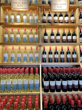 Shelves filled with rows of wine bottles, organized in an orderly fashion. The bottles have labels with blue and white designs, and the necks of the bottles are capped with red tops. Above the shelves, signs indicate different varieties and prices such as 'Tidal Bay' and 'Triumphe'. The shelves are wooden, creating a warm, inviting atmosphere.