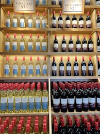 Shelves filled with rows of wine bottles, organized in an orderly fashion. The bottles have labels with blue and white designs, and the necks of the bottles are capped with red tops. Above the shelves, signs indicate different varieties and prices such as 'Tidal Bay' and 'Triumphe'. The shelves are wooden, creating a warm, inviting atmosphere.