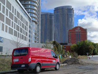 A red van is parked on a dirt road near a construction site, with modern high-rise buildings in the background. The sky is partly cloudy, and there is a red brick hotel with the word 'Delta' on it.