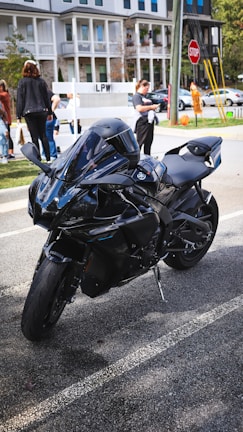 A sleek black motorcycle is parked on a paved street in an urban setting, with people walking and a red stop sign visible in the background. Three cars are parked along the curb, and several multi-story residential buildings are present nearby.