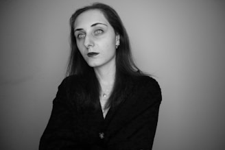 A person with long dark hair stands against a plain background. Their expression is neutral, and they are wearing a dark top. The image is in black and white, highlighting the person's facial features and creating a stark contrast with the background.