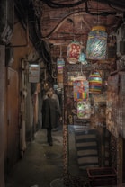 A narrow, dimly-lit alley adorned with colorful, artistic lanterns hanging from the ceiling. The walls are lined with various signs and a quaint establishment appears on the right. A couple of people walk away, creating a sense of mystery and exploration.