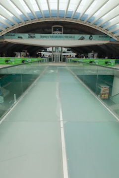 A modern architectural structure with a glass walkway leading towards a central area. Overhead, there is an advertisement banner for Samsung products. The ceiling features a grid-like pattern with skylights allowing natural light to enter. The sides of the walkway are lined with glass railings, and there are green advertisements suggesting promotions.