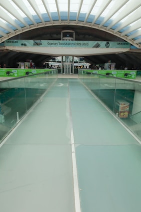 A modern architectural structure with a glass walkway leading towards a central area. Overhead, there is an advertisement banner for Samsung products. The ceiling features a grid-like pattern with skylights allowing natural light to enter. The sides of the walkway are lined with glass railings, and there are green advertisements suggesting promotions.