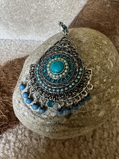An intricately designed piece of jewelry, featuring a teardrop shape adorned with concentric circles of small turquoise beads and metal filigree. It's set against a textured stone background that contrasts with the detailed design.