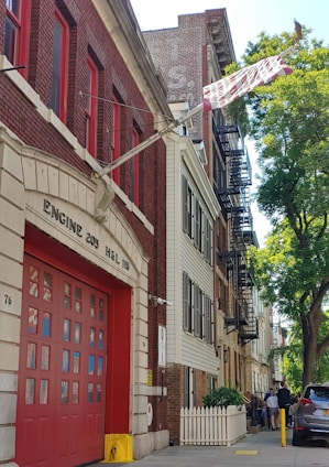 A red brick fire station with large garage doors is positioned on a residential street. The building is labeled 'Engine 205 H&L 118.' Beside it are multi-story residential buildings with fire escapes. A group of people stands on the sidewalk near a white picket fence, and there is a tree providing shade.
