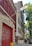 A red brick fire station with large garage doors is positioned on a residential street. The building is labeled 'Engine 205 H&L 118.' Beside it are multi-story residential buildings with fire escapes. A group of people stands on the sidewalk near a white picket fence, and there is a tree providing shade.
